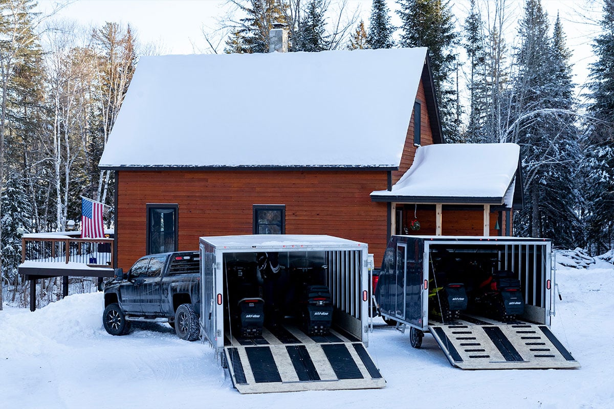 Crossover & Crossover 2.0 aluminum snow trailers parked at a cabin in winter, ramp doors open to unload