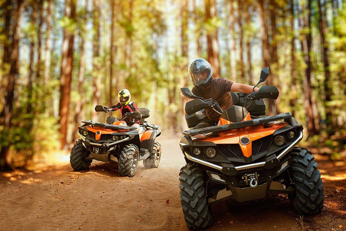 Two ATV riders on a dirt trail through a forest