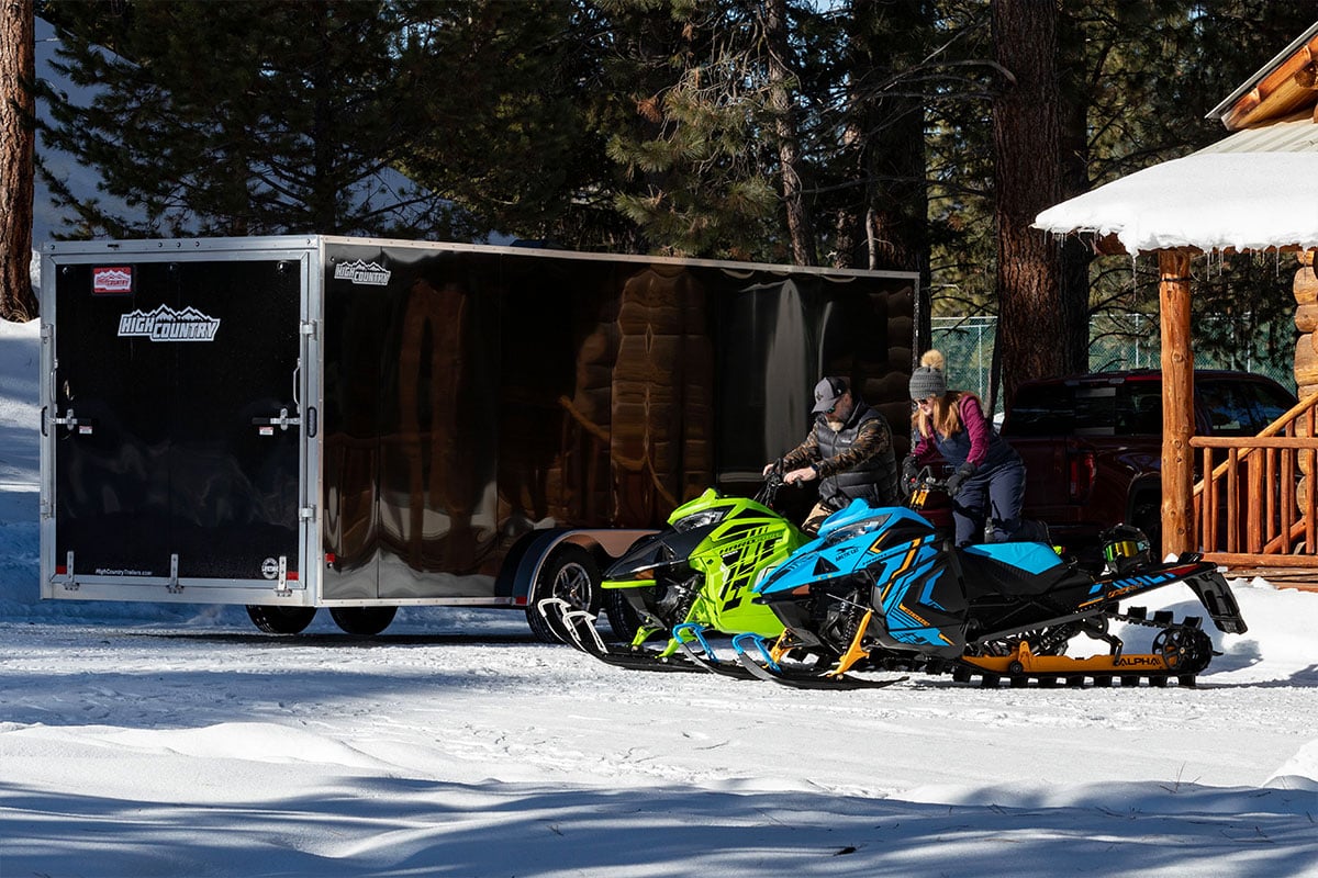 Black aluminum Inline snow trailer and two snowmobiles parked outside a log cabin in winter