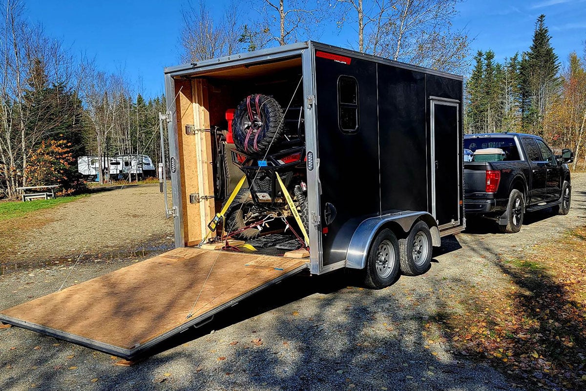 Black enclosed UTV/ATV trailer with rear ramp door open at a campsite. Custom interior setup for camping.