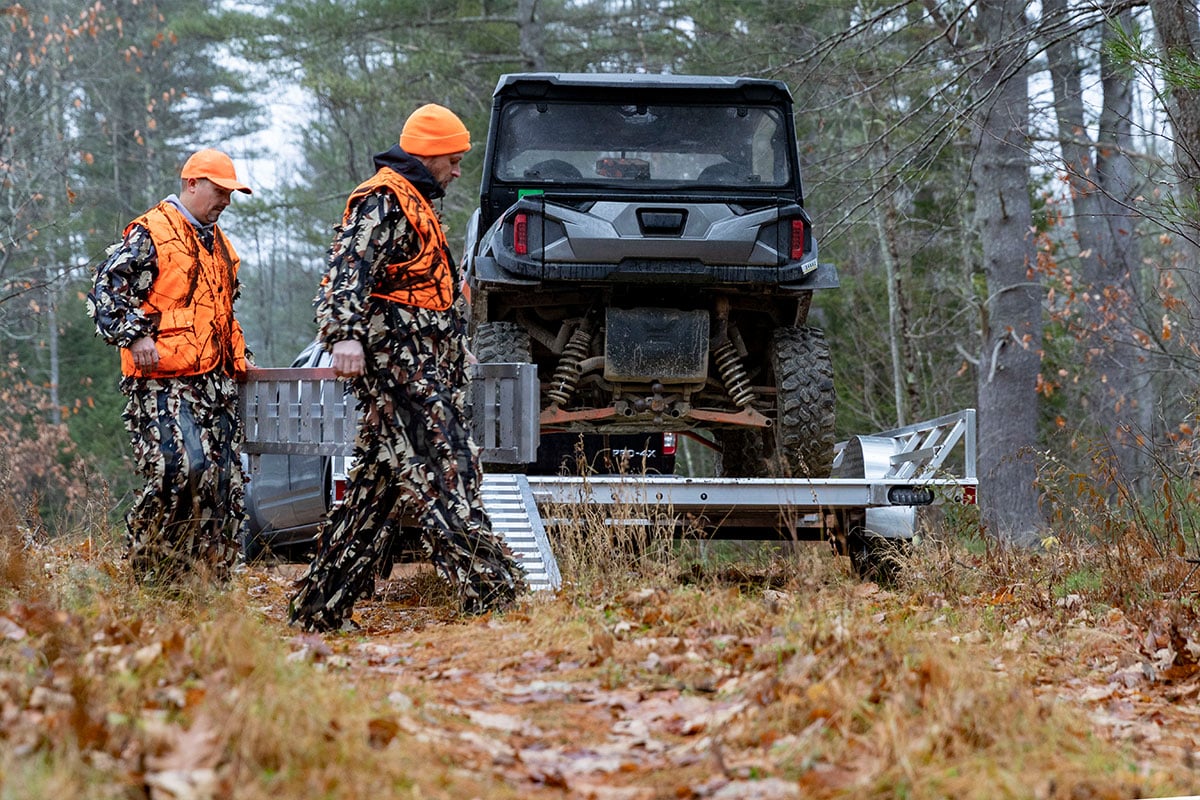 Open ALCOM trailer parked in the woods; two men are setting up the portable ramps to unload.