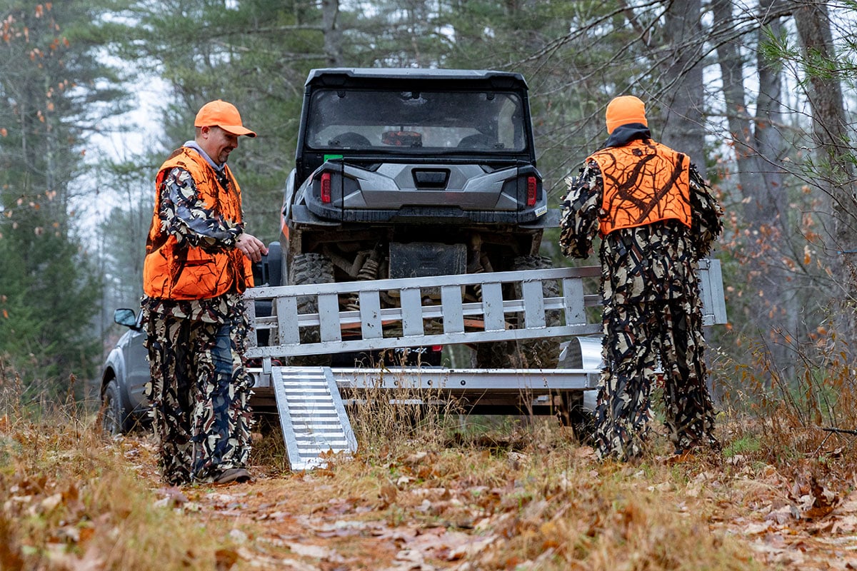 Two men dressed in camo and safety orange placing ramps on an open ALCOM trailer.