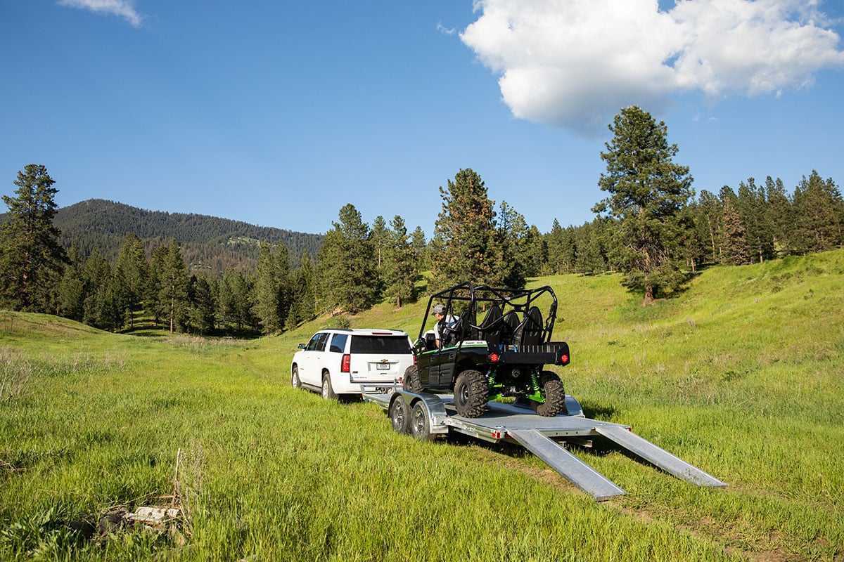 ATV loaded onto an open aluminum ALCOM trailer with stowaway ramps, parked in a beautiful meadow.