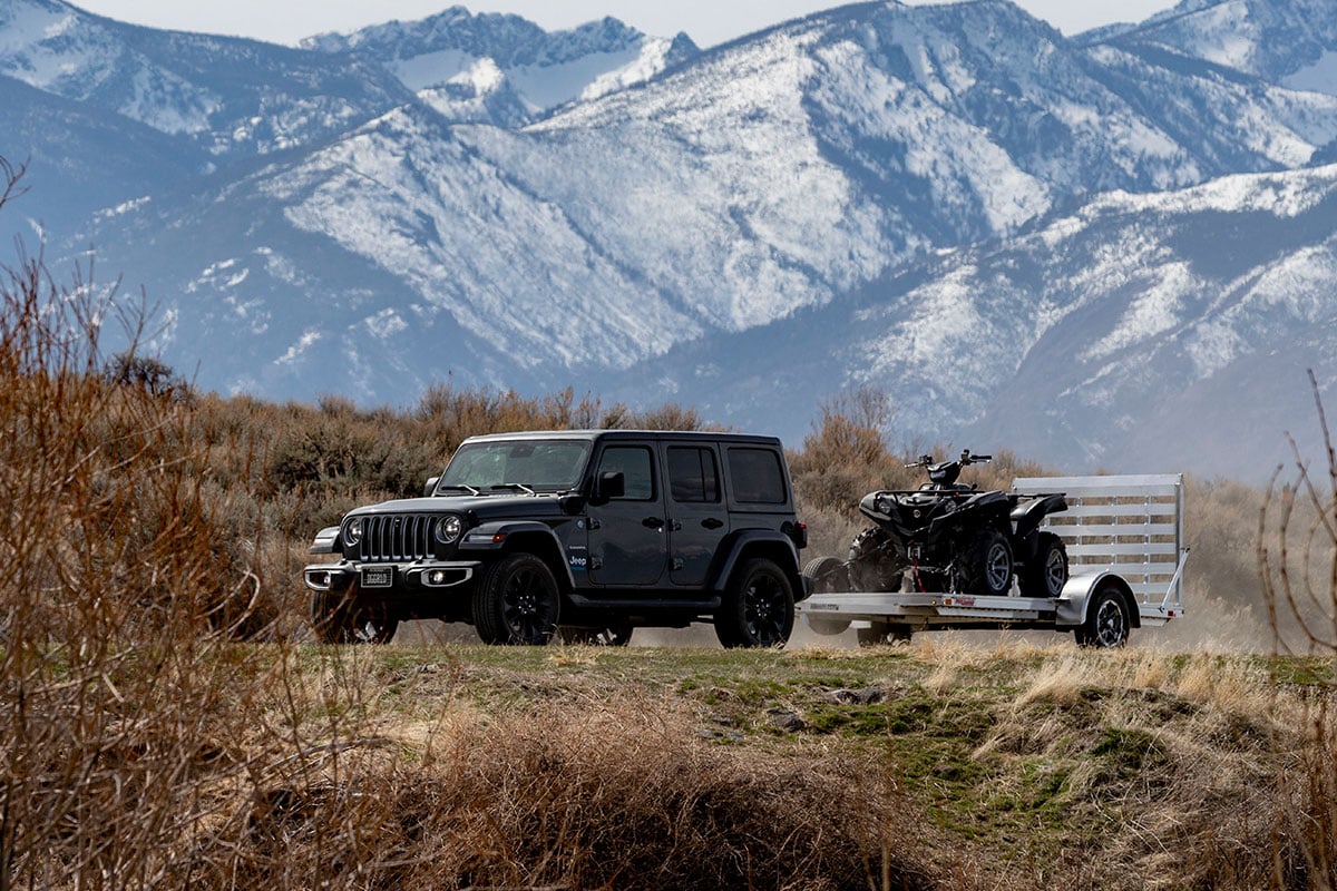 Open ATV trailer pulled by a Jeep in front of a mountain range