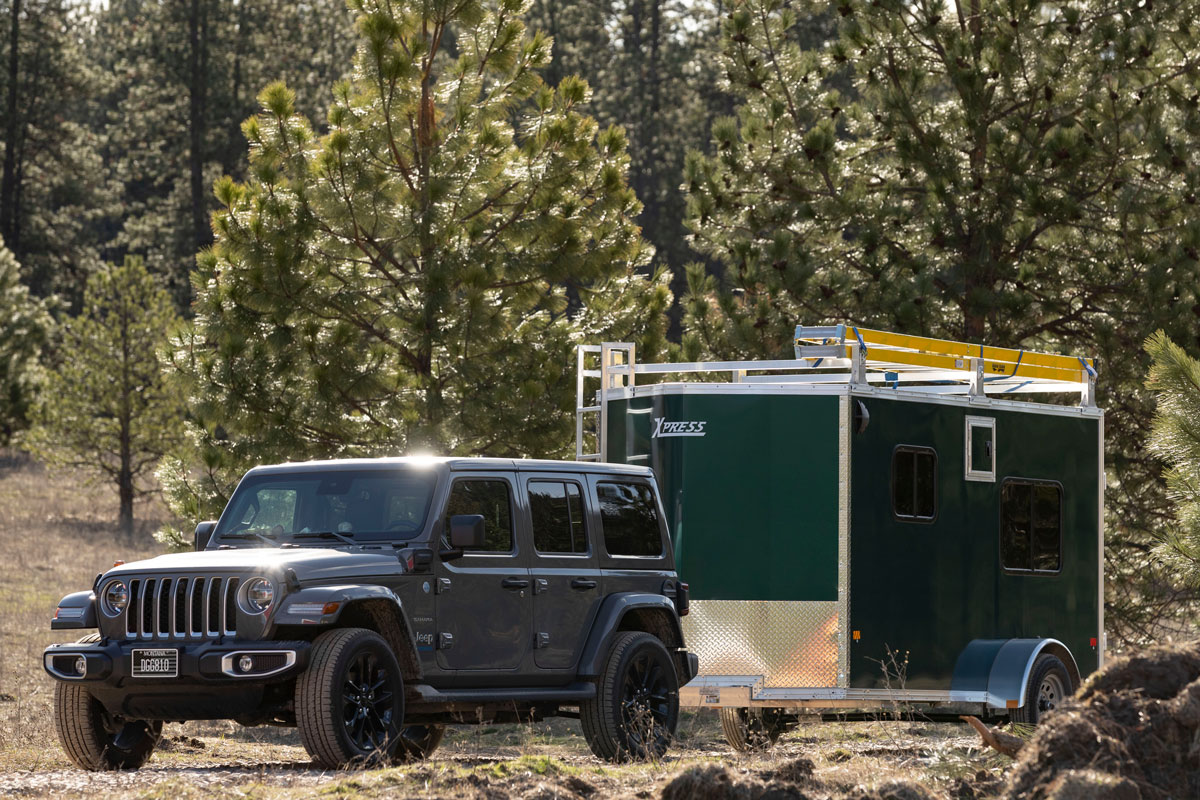 A 5' wide green enclosed cargo trailer hauled by a Jeep, featuring multiple windows, a ladder to the roof, and multiple rooftop ladder racks