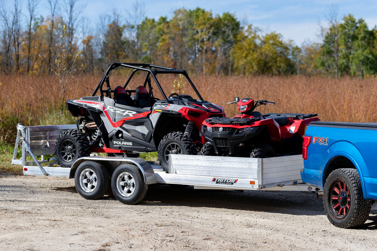 An ALCOM open UTV trailer with two machines parked on a gravel lot