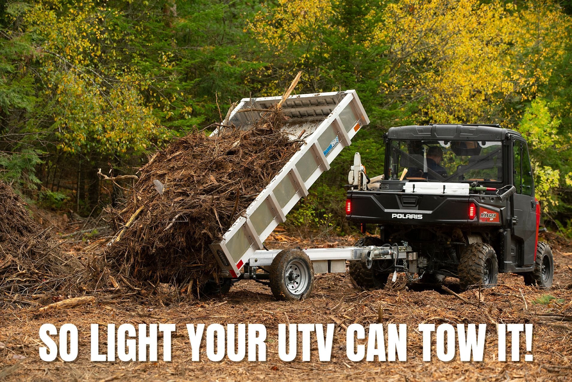 A side-by-side vehicle tows an ALCOM residential grade aluminum dump trailer, shown dumping brush in a Maine autumn yardwork scene