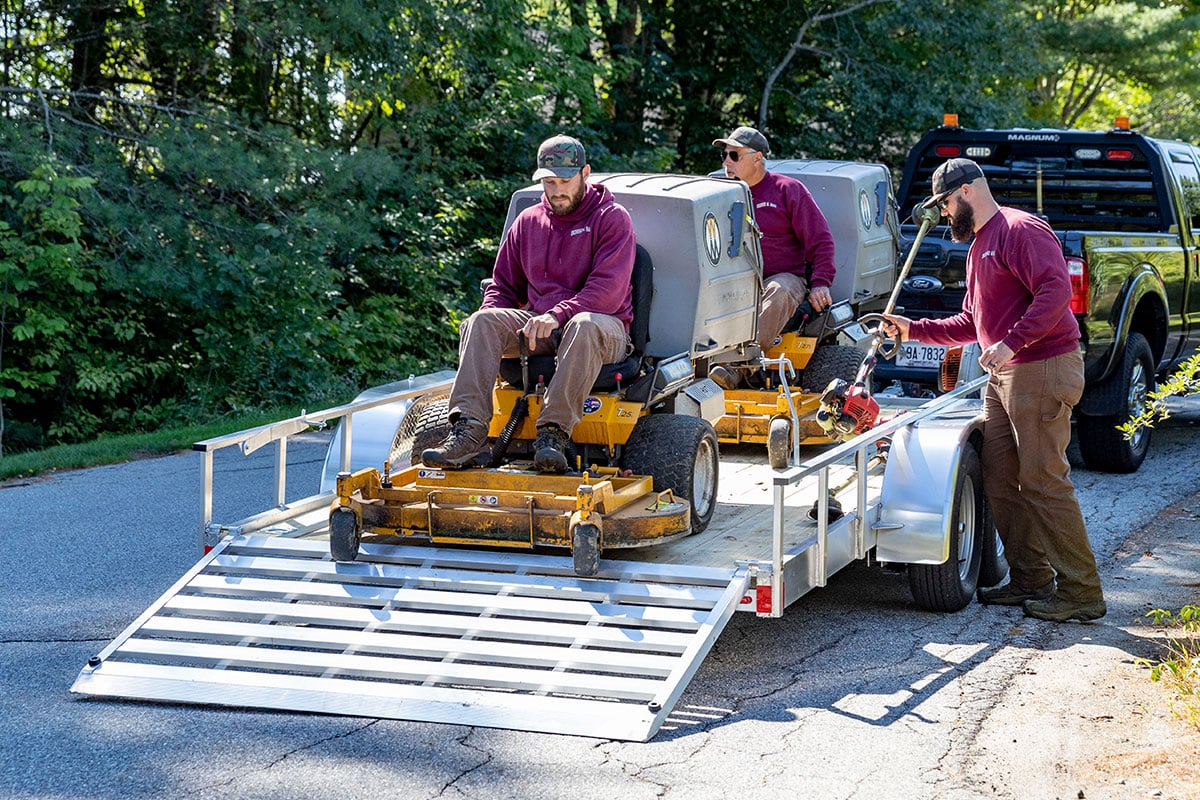 Open aluminum utility trailer from ALCOM with two people driving lawn equipment down rear ramp