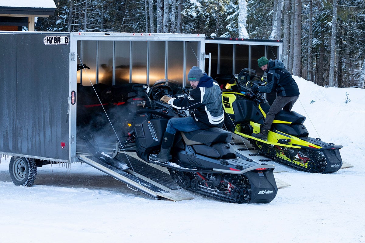 Two enclosed ALCOM snow trailers; two people are unloading snowmobiles on the rear ramps.
