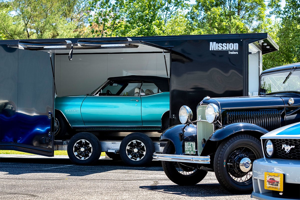 Side view of a sleek black Mission enclosed aluminum car hauler, with a blue convertible visible through the open Elite Escape Door.