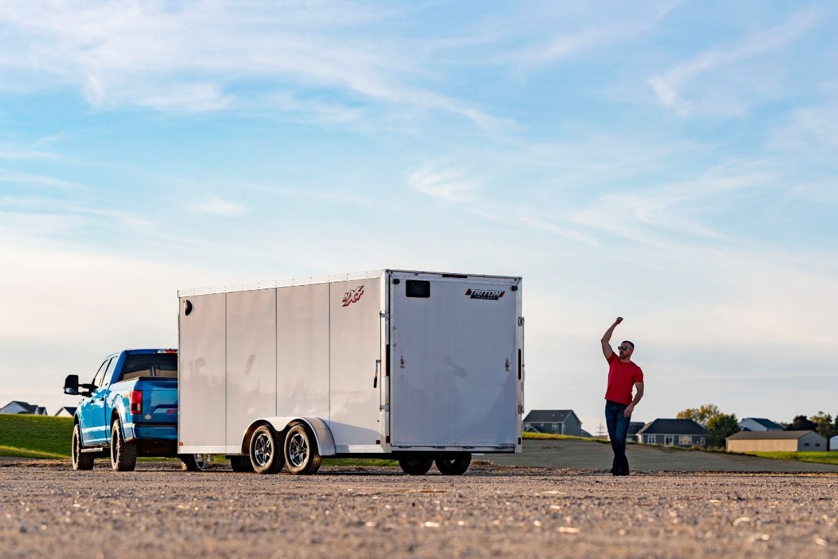 A white enclosed aluminum cargo trailer by ALCOM hitched to a blue truck parked in a gravel field; a man has just closed the spring assisted rear ramp door 