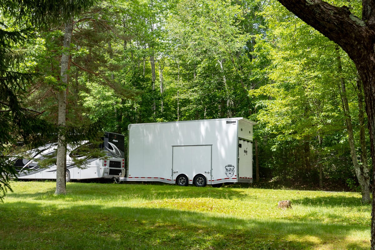 White enclosed Pinnacle Stacker car hauler trailer hitched to an RV driving through trees on a sunny day