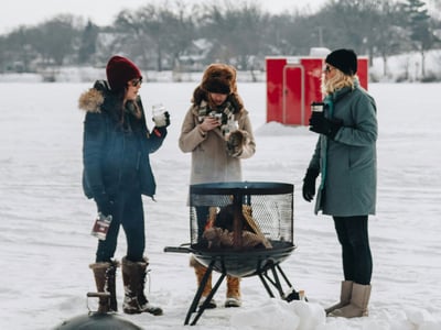 Friends standing around a fire pit on frozen lake with red aluminum ice shack in the background