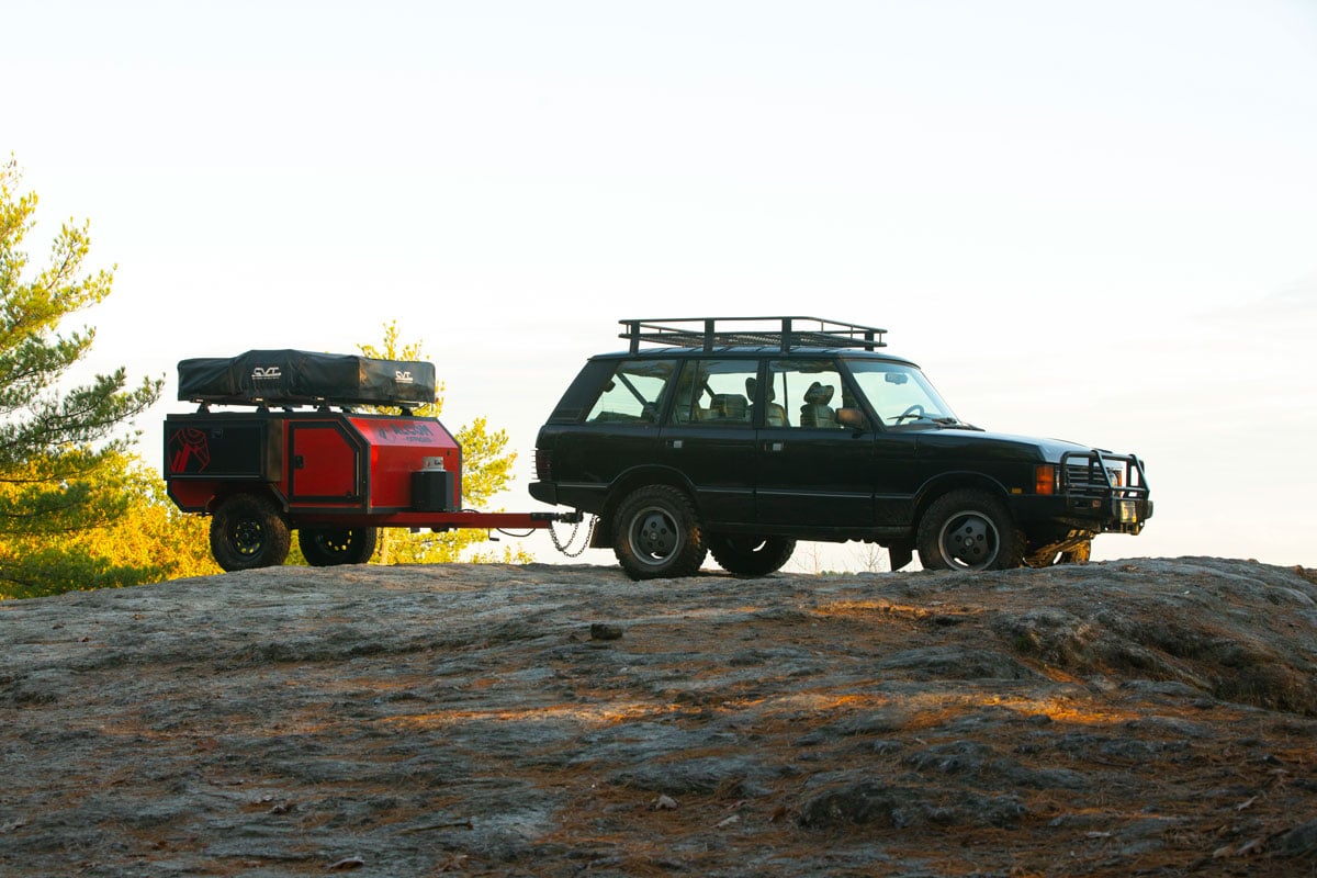A lightweight red Spur offroad trailer by AO Trailers towed by an SUV to the top of a rocky hill overlooking the sunset