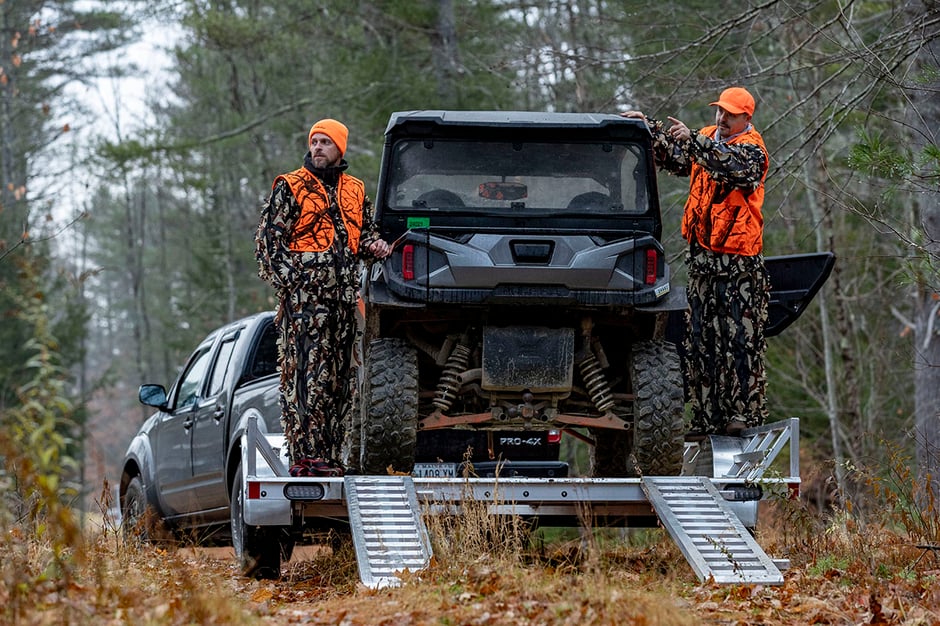 Two men in camo and safety orange standing on the trailer by a UTV, looking into the woods