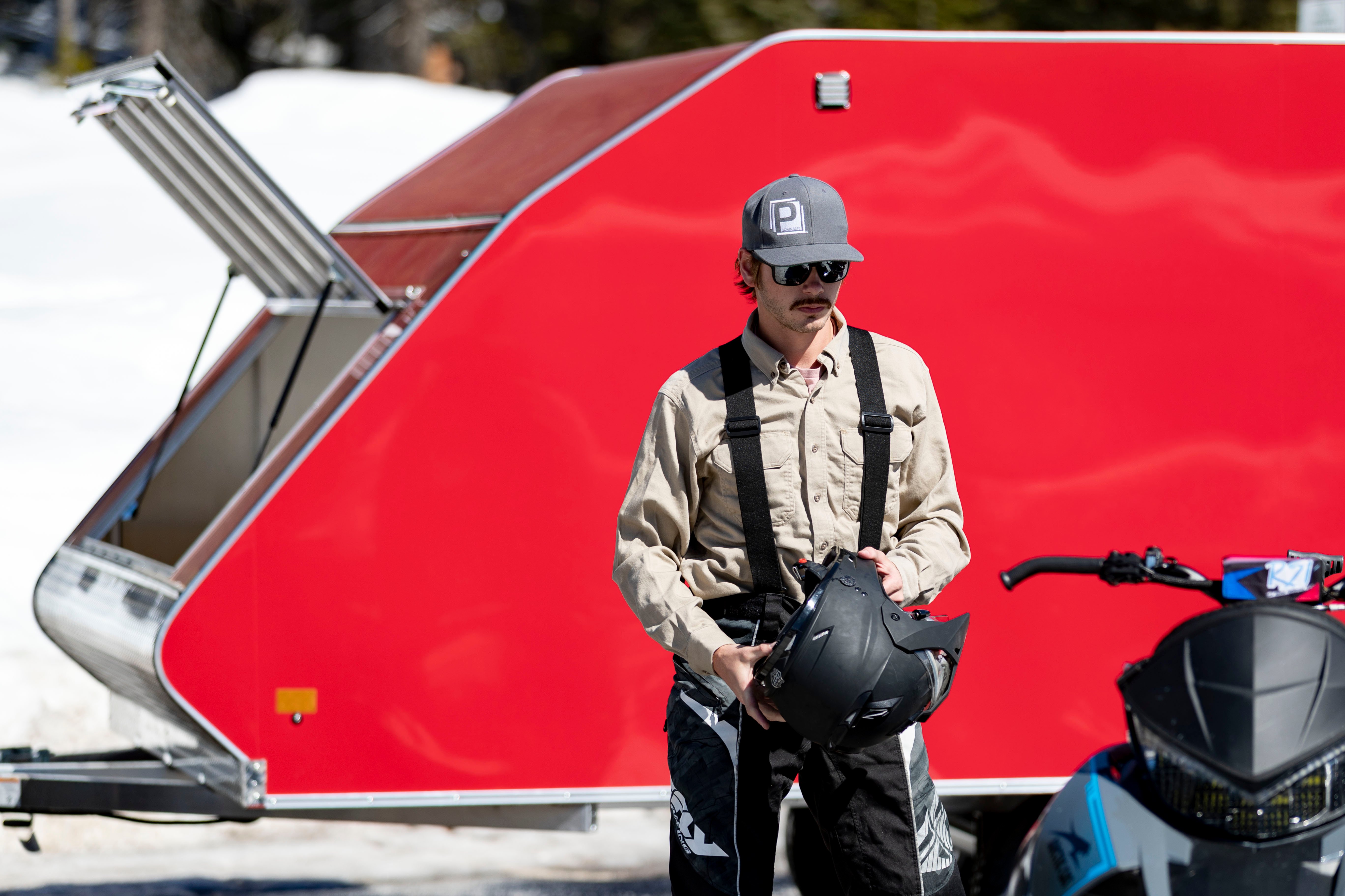 A snowmobiler gets ready to ride in front of a red enclosed Crossover trailer