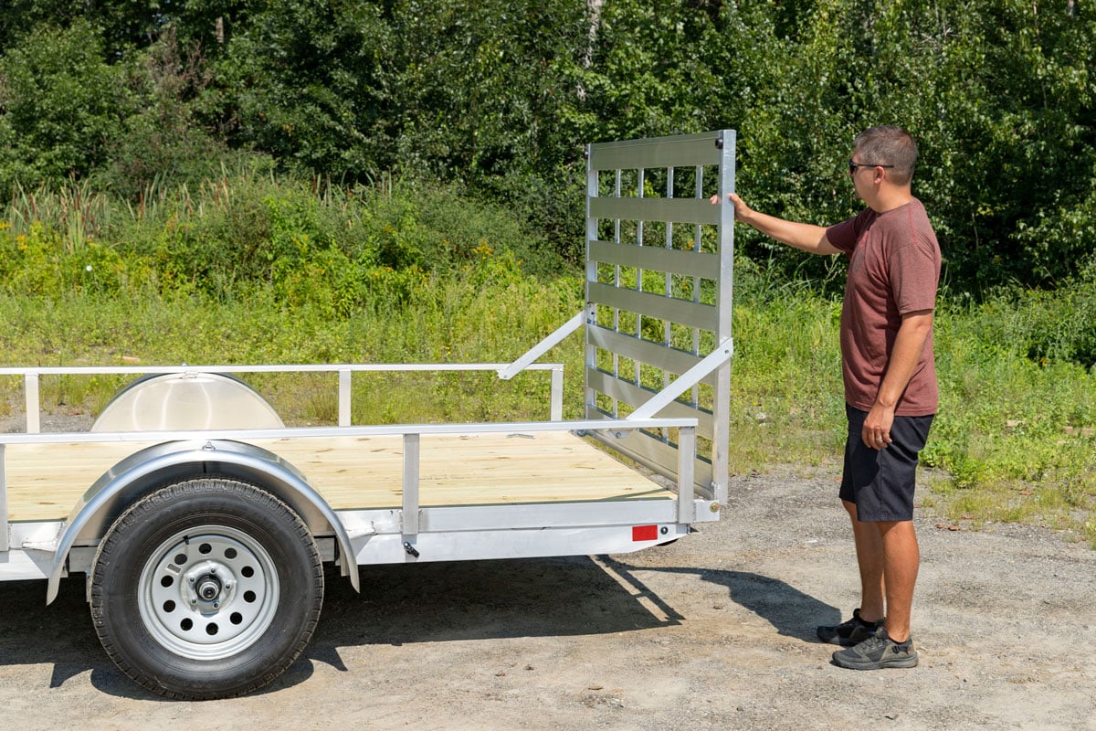 A versatile open aluminum utility trailer with wood deck and side rail kit; a man gets ready to lower the rear ramp on a summer day