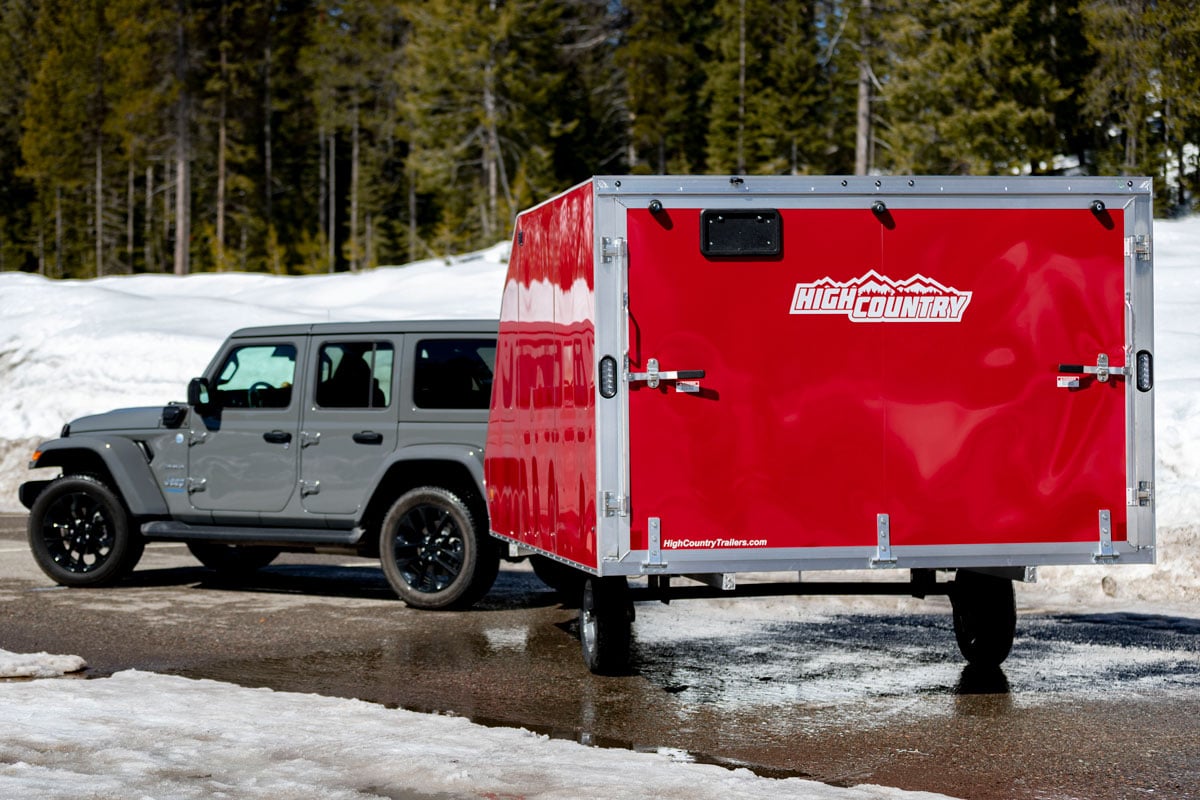 A red High Country crossover snowmobile trailer hitched to a gray Jeep parked on a paved lot