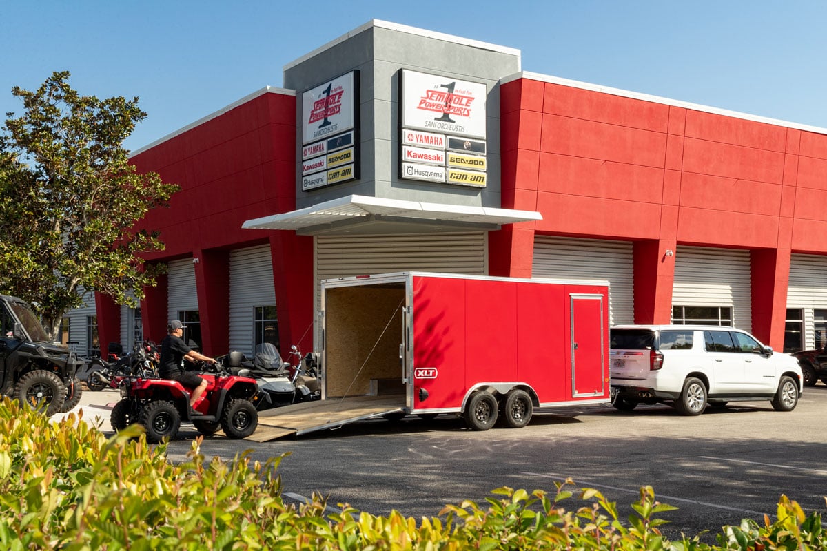 A man drives an ATV up the rear ramp of a red XLT Pro Series car hauler by ALCOM outside a power sports dealership