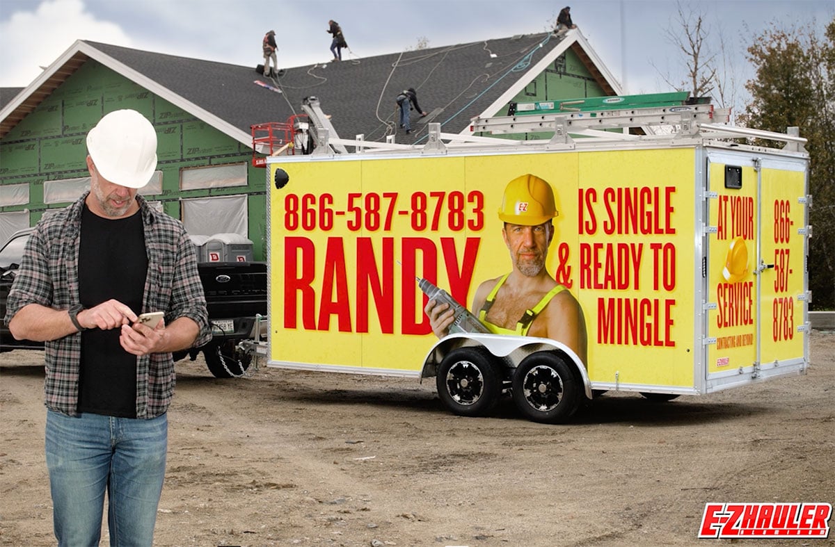 A contractor answers a call from a new customer while standing on a jobsite with his humorously wrapped ALCOM aluminum trailer advertising handyman services