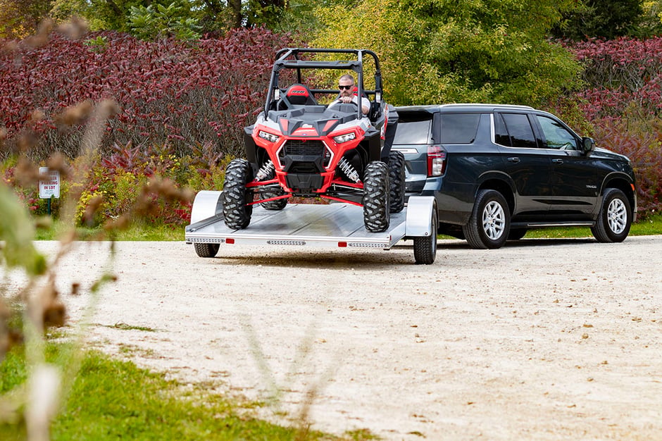 A man unloads a UTV from a tilting aluminum trailer hitched to an SUV parked on a gravel lot