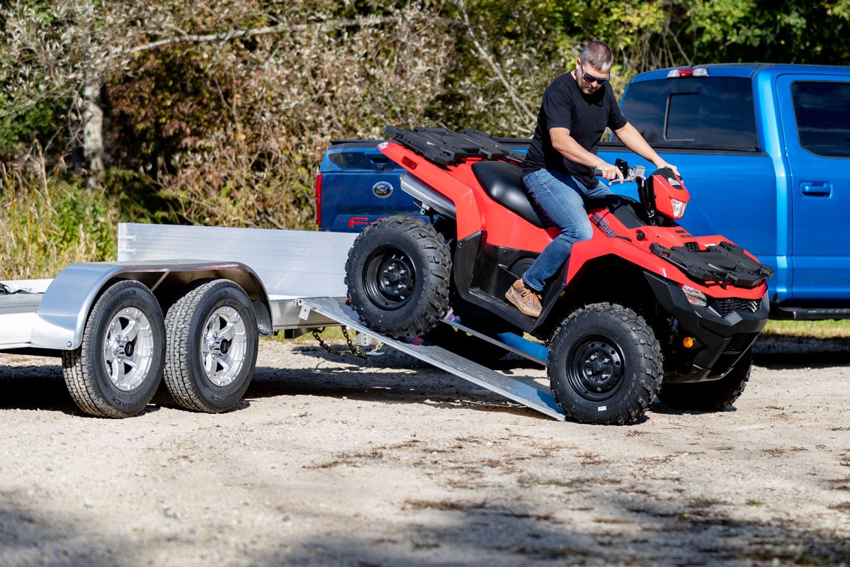A man unloading an ATV from an open aluminum trailer using ALCOM's side rail kit on a sunny autumn day