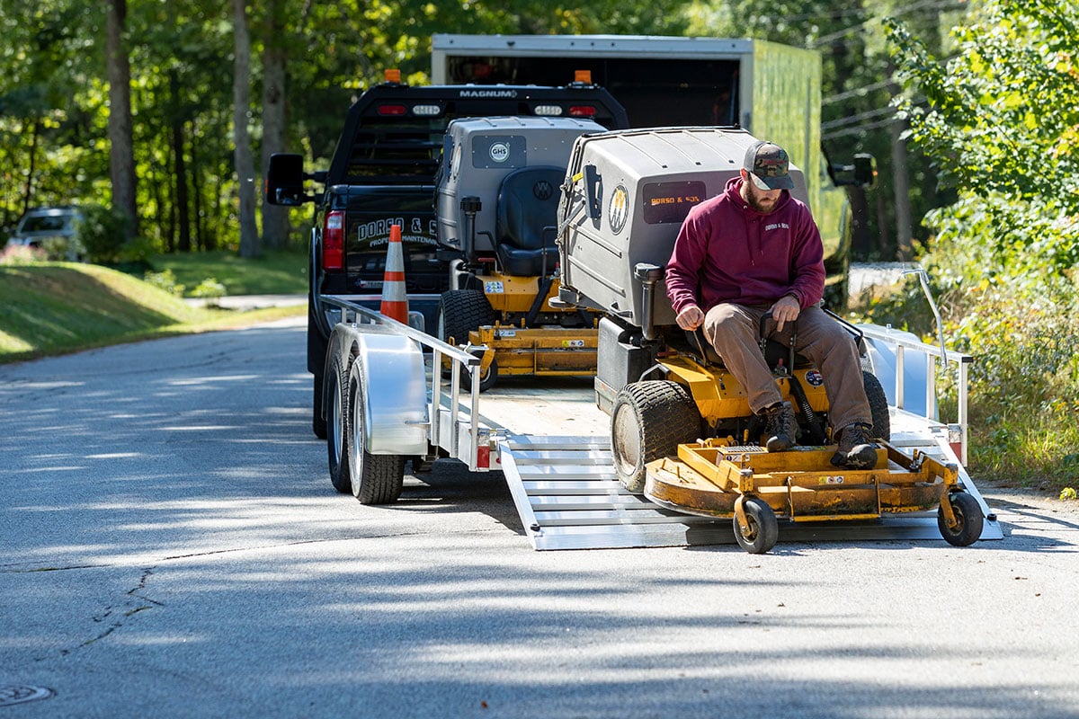 A landscaper unloading a riding lawnmower from an open wood deck utility trailer by ALCOM