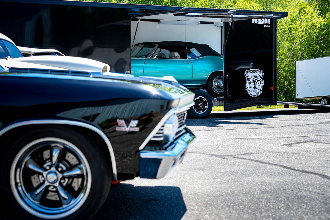 A sleek black enclosed aluminum Pinnacle car hauler trailer parked with shiny classic cars on a sunny day.