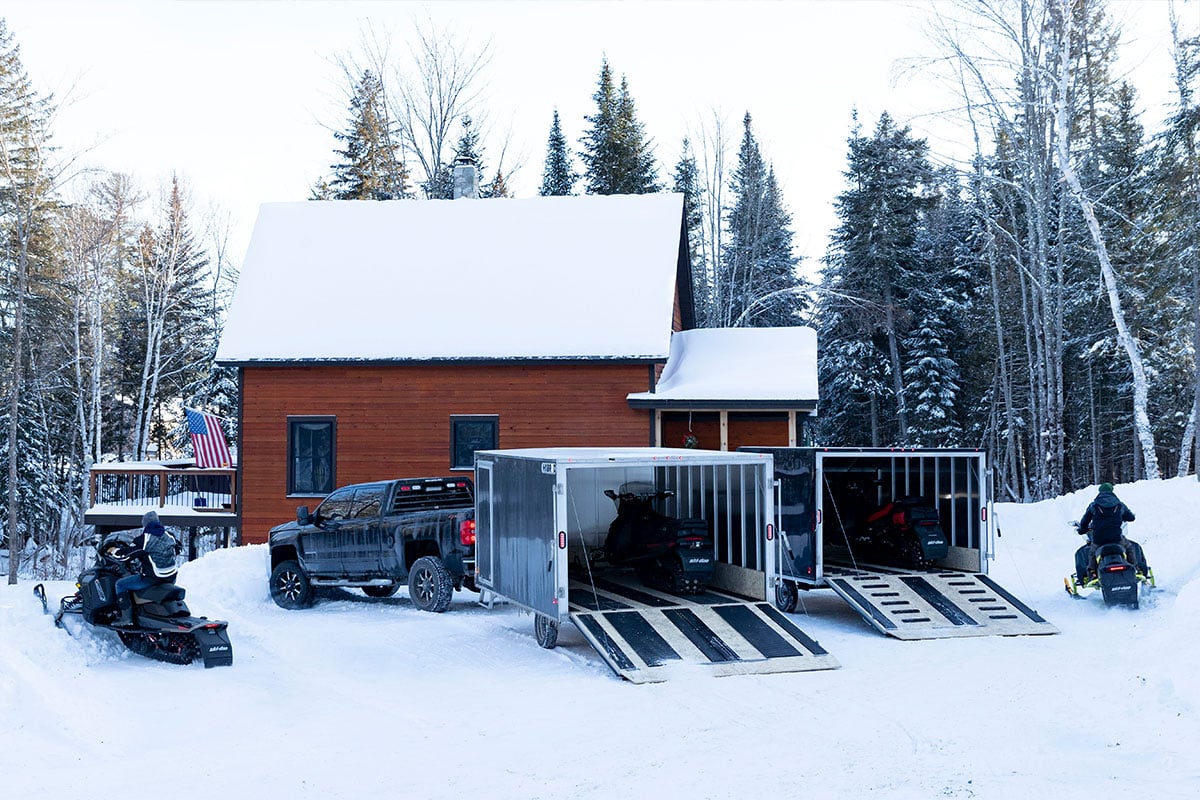 Two enclosed aluminum crossover snow trailers by ALCOM parked with rear ramp doors open and snowmobiles driving away