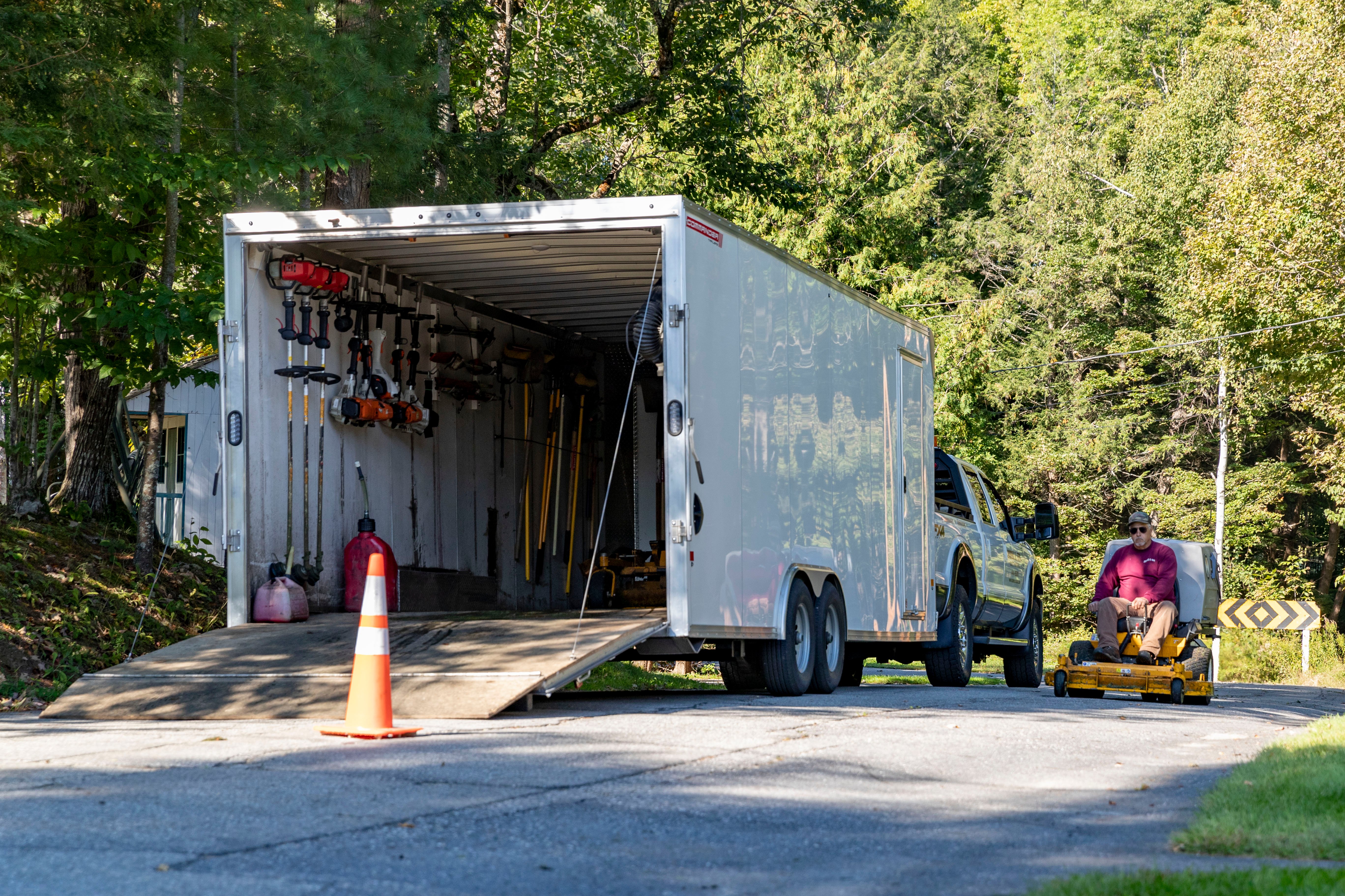 Large enclosed aluminum cargo trailer with rear ramp door loaded with landscaping tools and equipment.