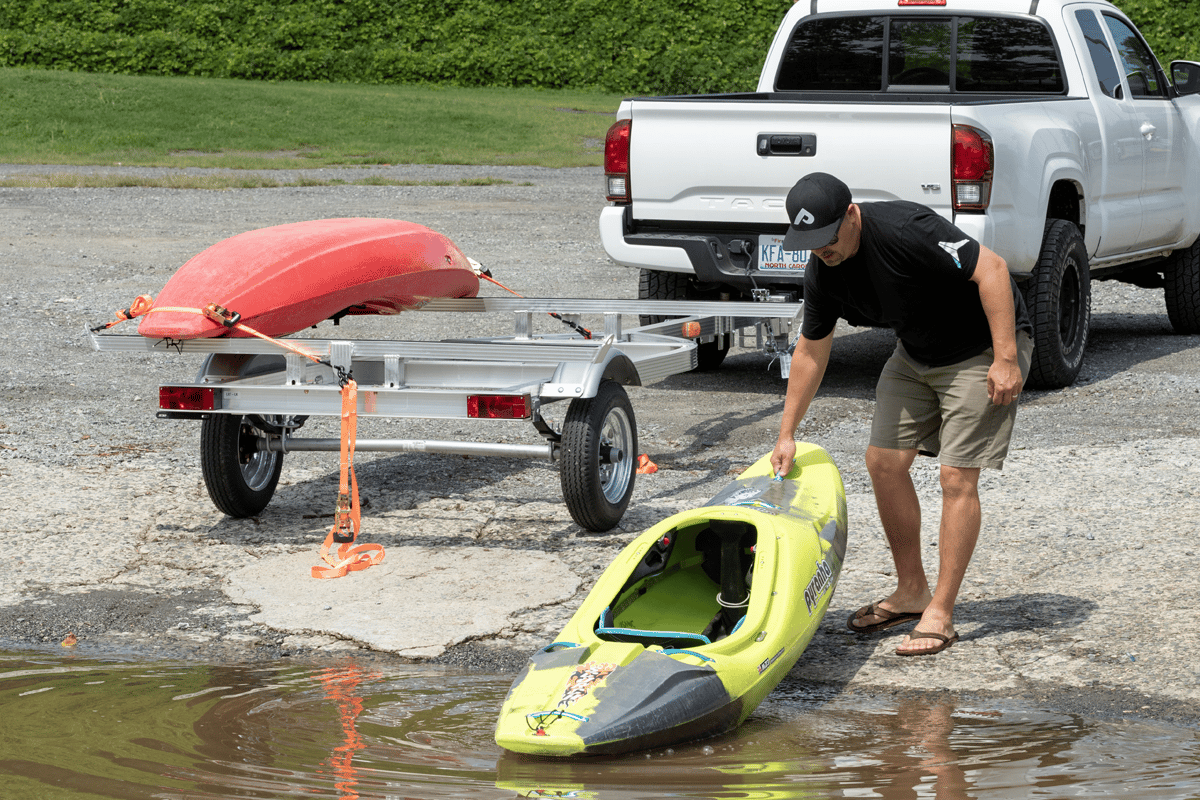A man pulling a kayak out of the water on a boat ramp; one kayak is already secured to his two-place aluminum kayak trailer