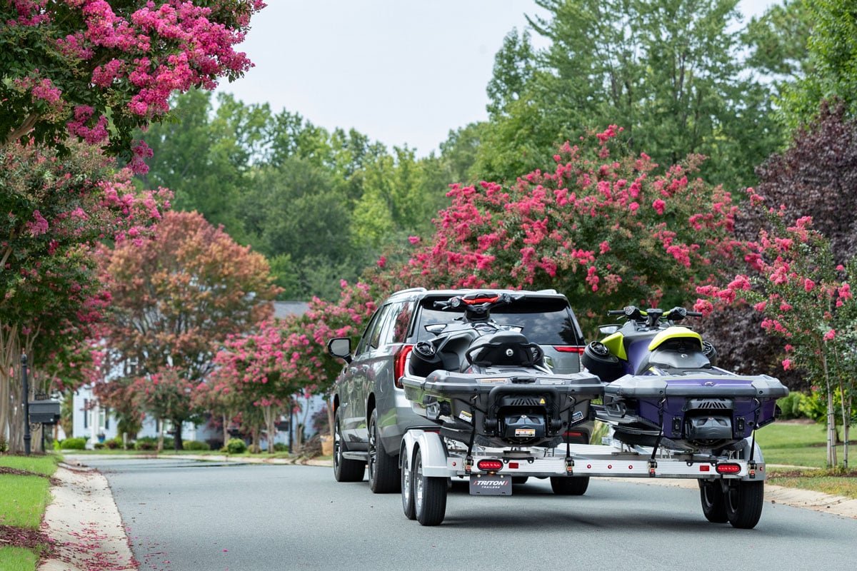 Silver SUV towing a Triton aluminum two-place PWC trailer through a scenic neighborhood with pink flowering trees. 