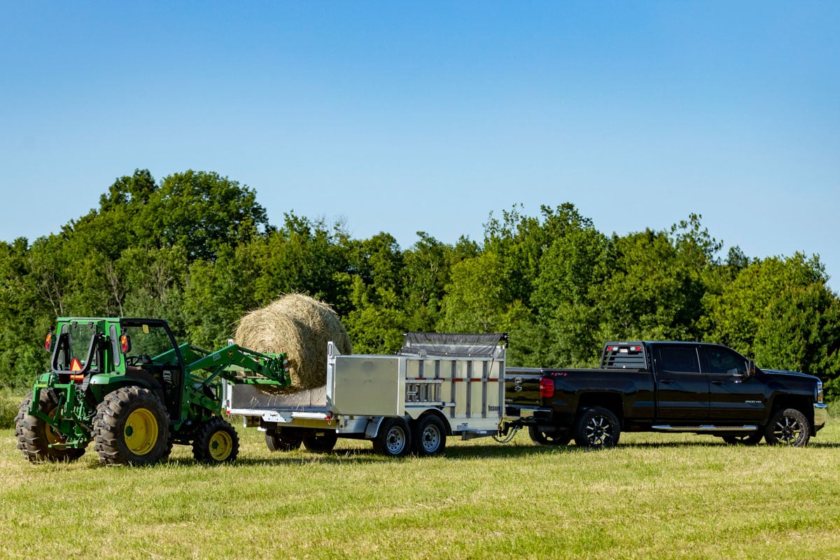 A tractor loading a large hay bale into a Mission Trailers commercial aluminum dump trailer.
