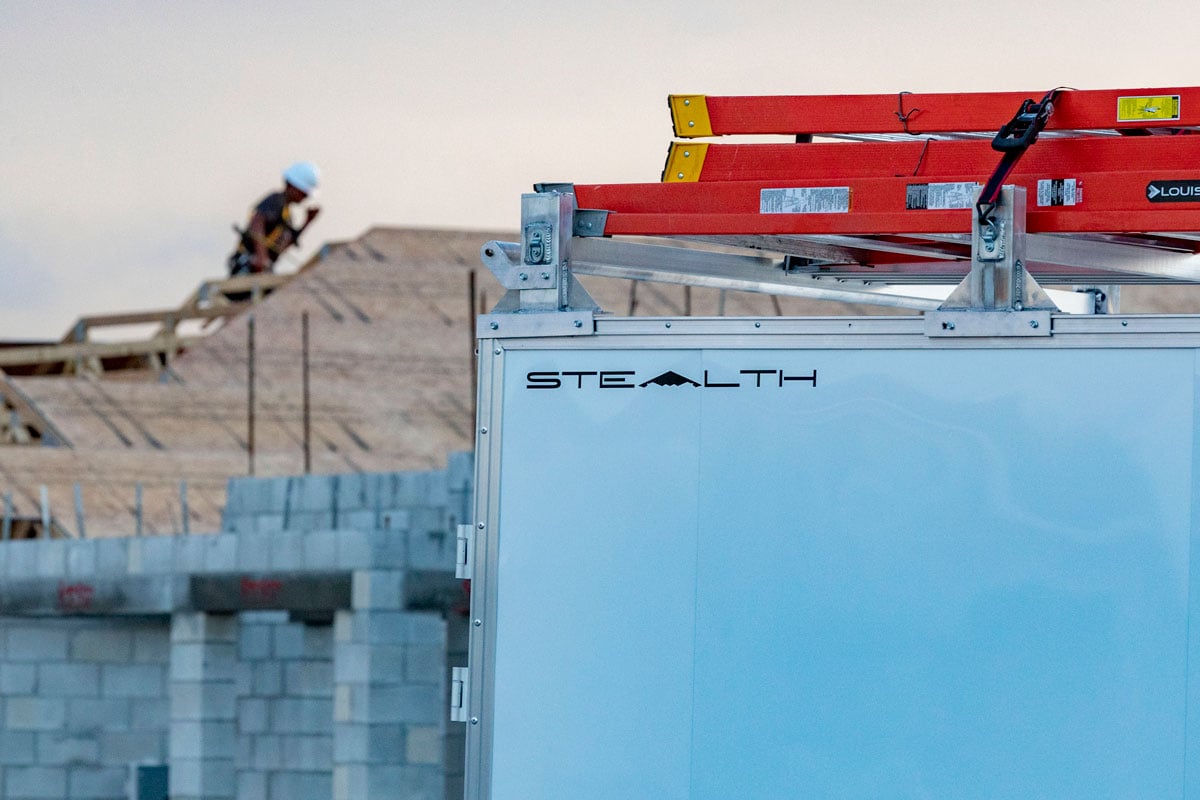A white Stealth enclosed Ultimate Contractor Package at a jobsite with ladders on rack and worker on the roof behind the trailer