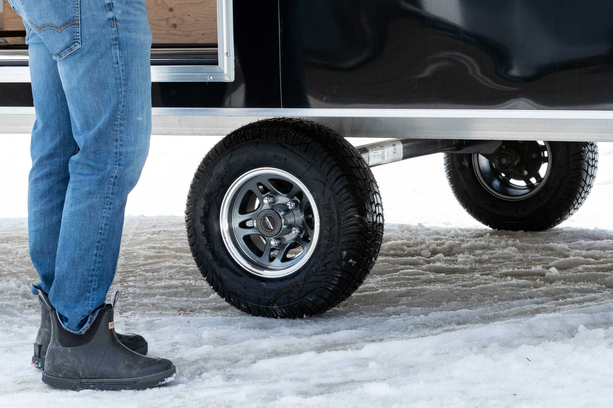 Close view of tires on an ALCOM aluminum snowmobile trailer and a man's feet standing beside it