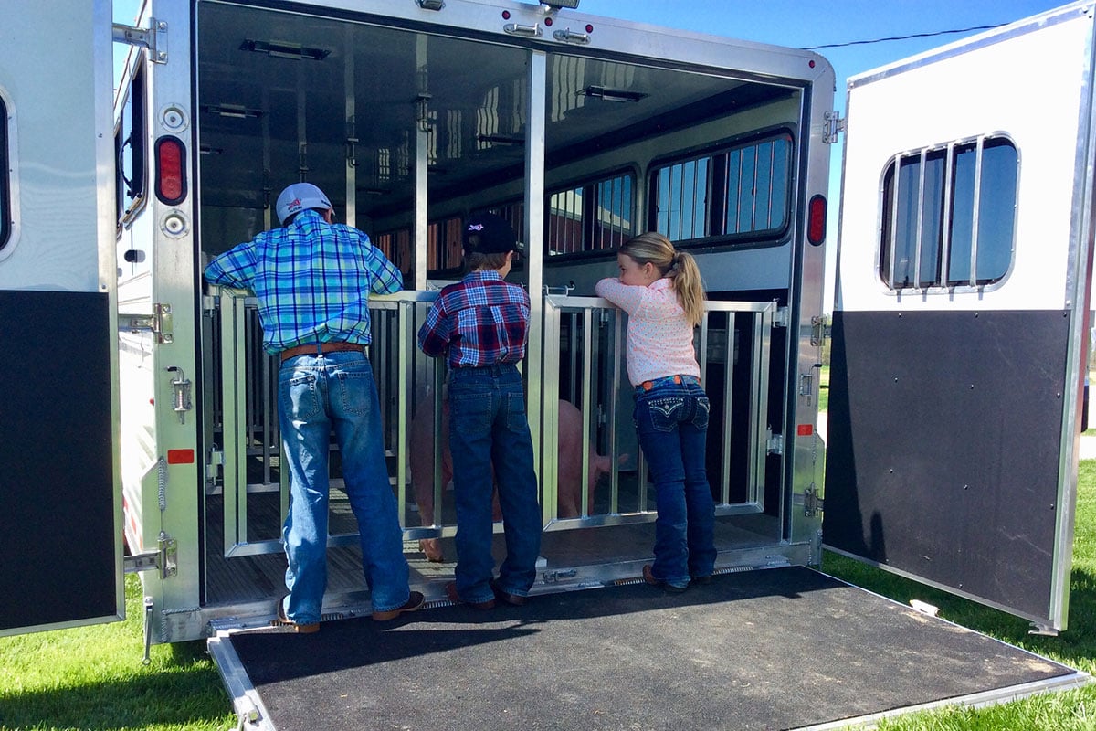 Three young farmers in training stand at the open loading end of a Frontier Stock LowPro trailer with pigs loaded safely into adjustable stalls.