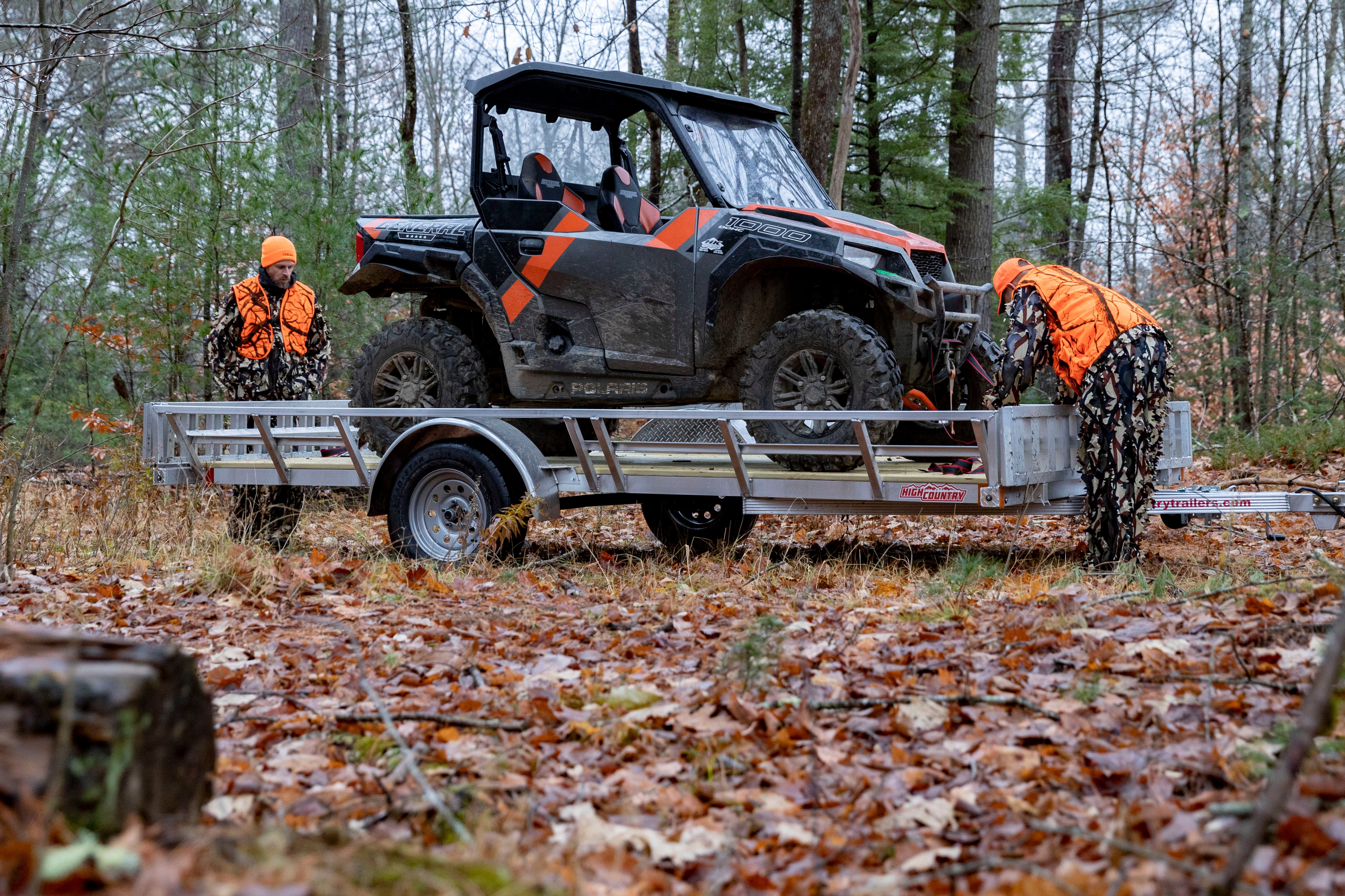 Two men in camo suits and safety orange vests and hats prepare to unload a UTV from an aluminum UTV trailer parked on a trail during autumn