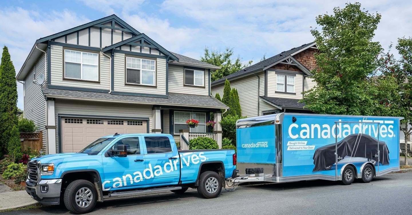 A custom wrapped blue Canada Drives pickup truck and Mission Car Hauler trailer parked in front of townhomes; PC Canada Drives via Mission FB