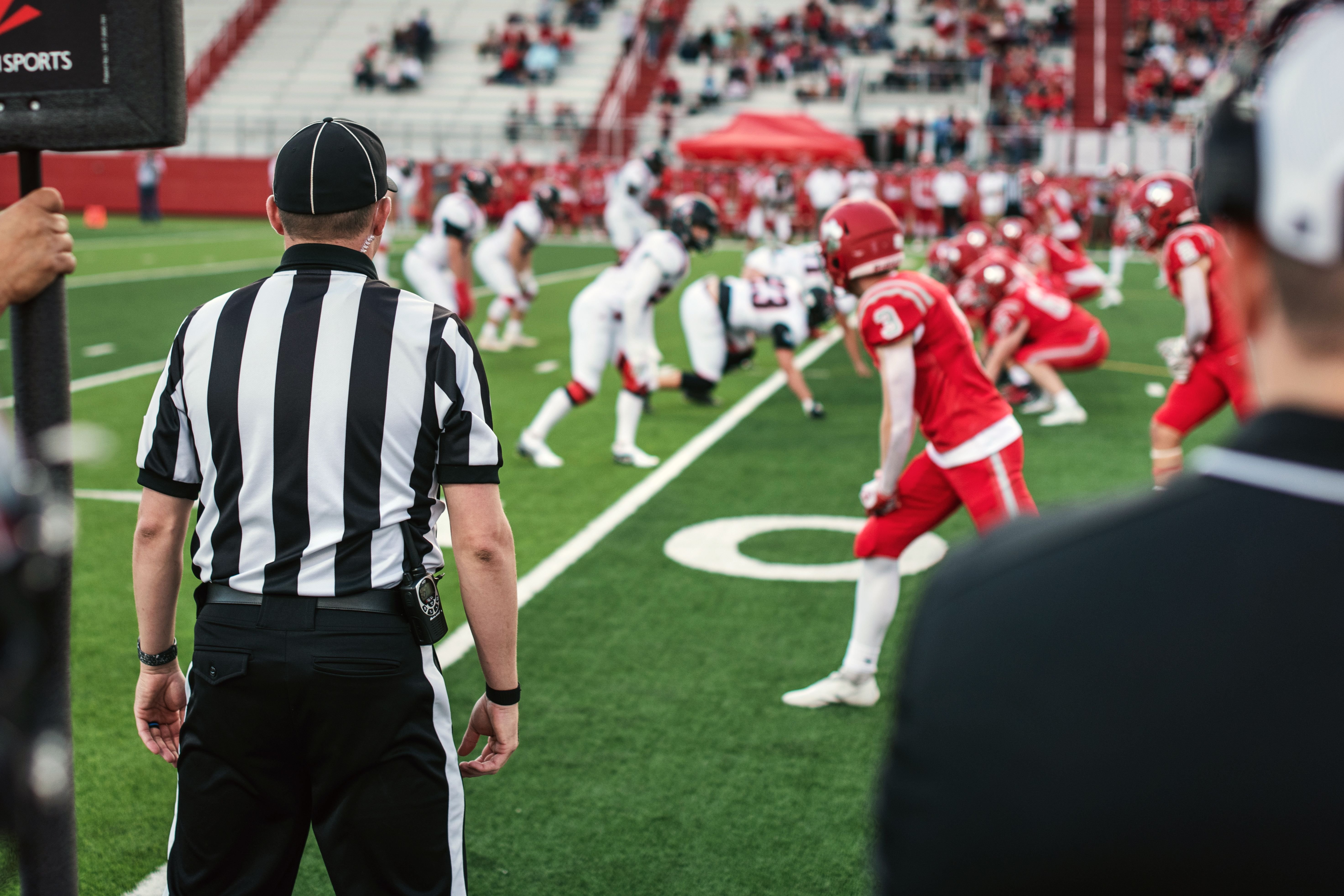 Referees and coaches on the sidelines of a high school football game with players lined up facing each other