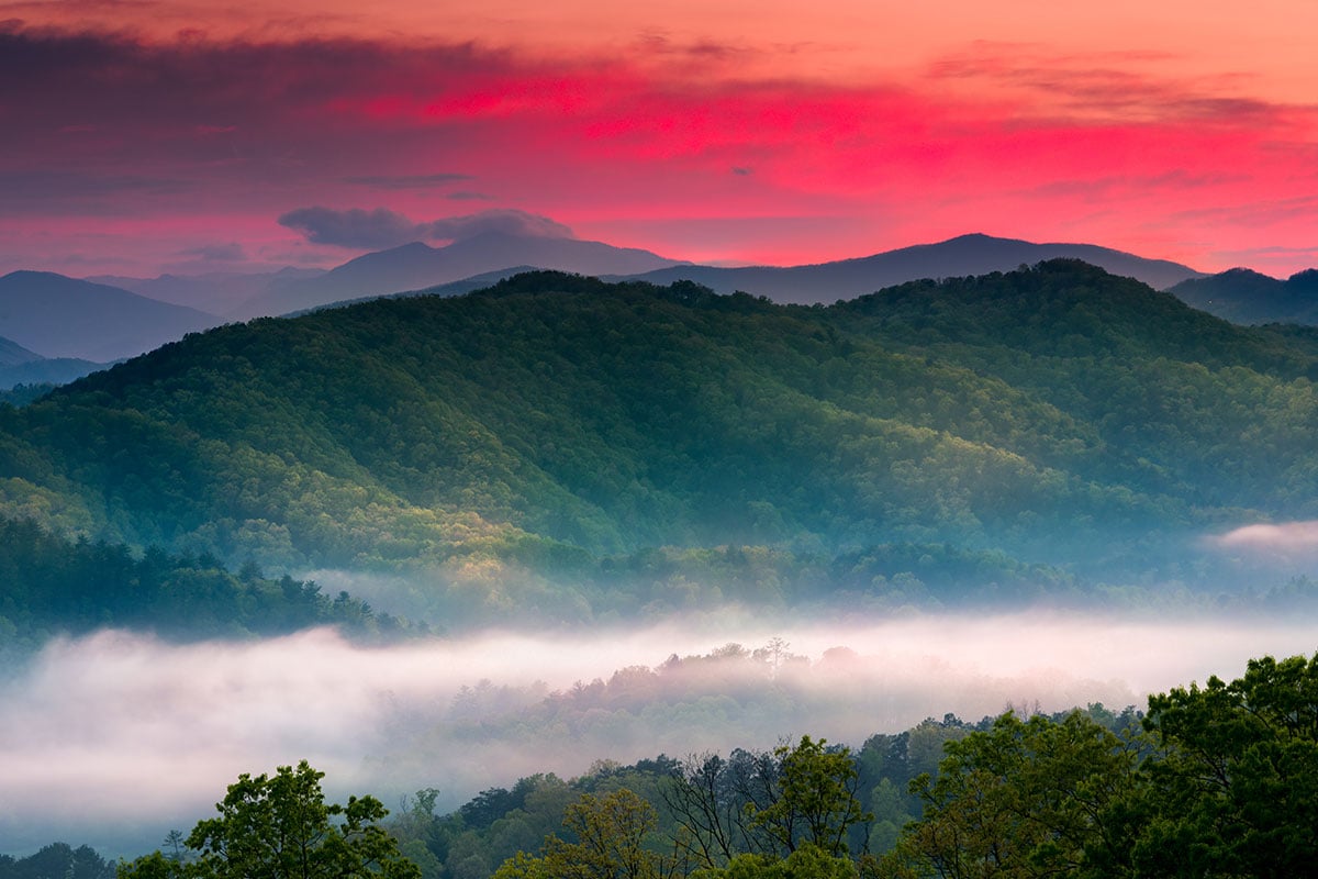 Scenic sunset view of the Great Smoky Mountains showing layered trees, fog, green mountains and a red sunset sky