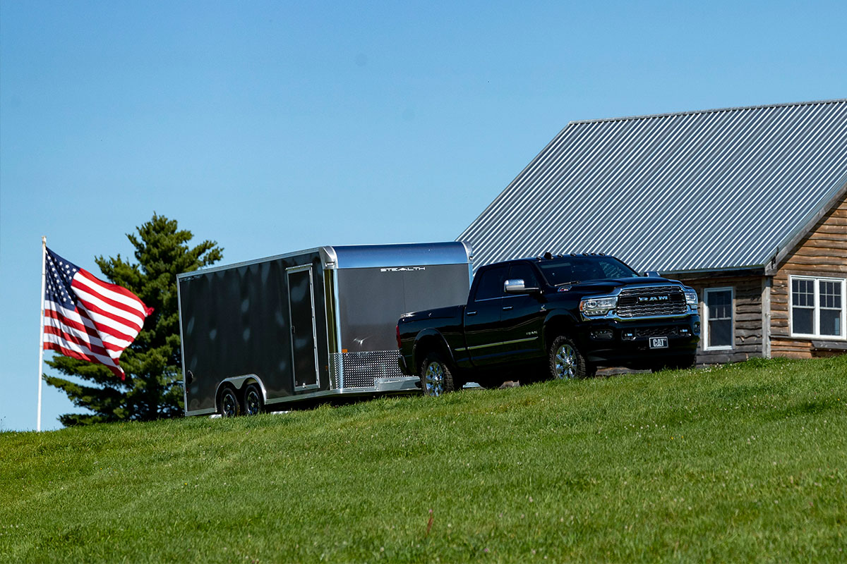 Enclosed aluminum car hauler by ALCOM, parked with an American flag and barn in the background.