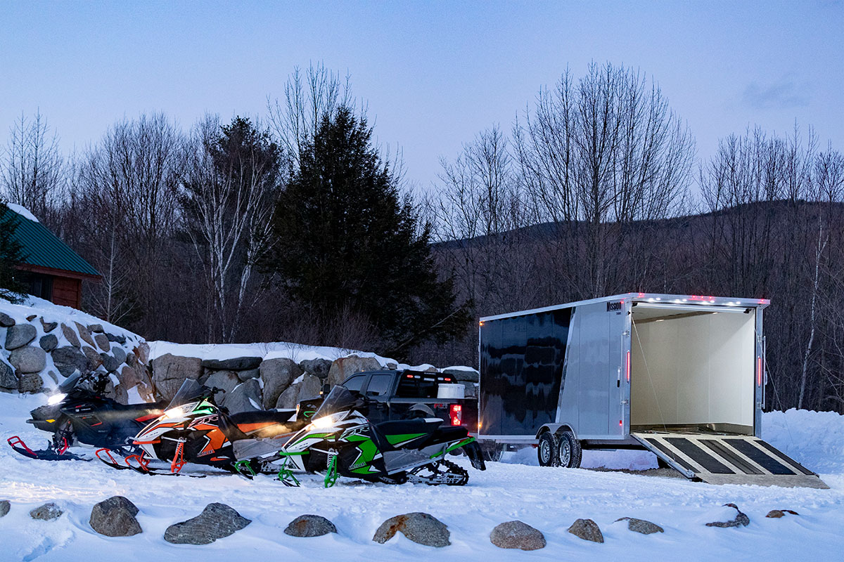 Enclosed ALCOM snow trailer parked with several snowmobiles, showing exterior ramp lights on the trailer.