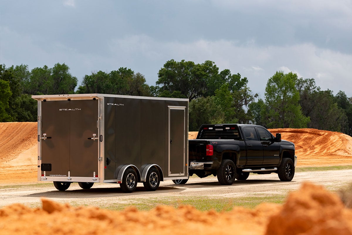 A black pickup truck tows a dark grey enclosed aluminum cargo hauler with a side access door.