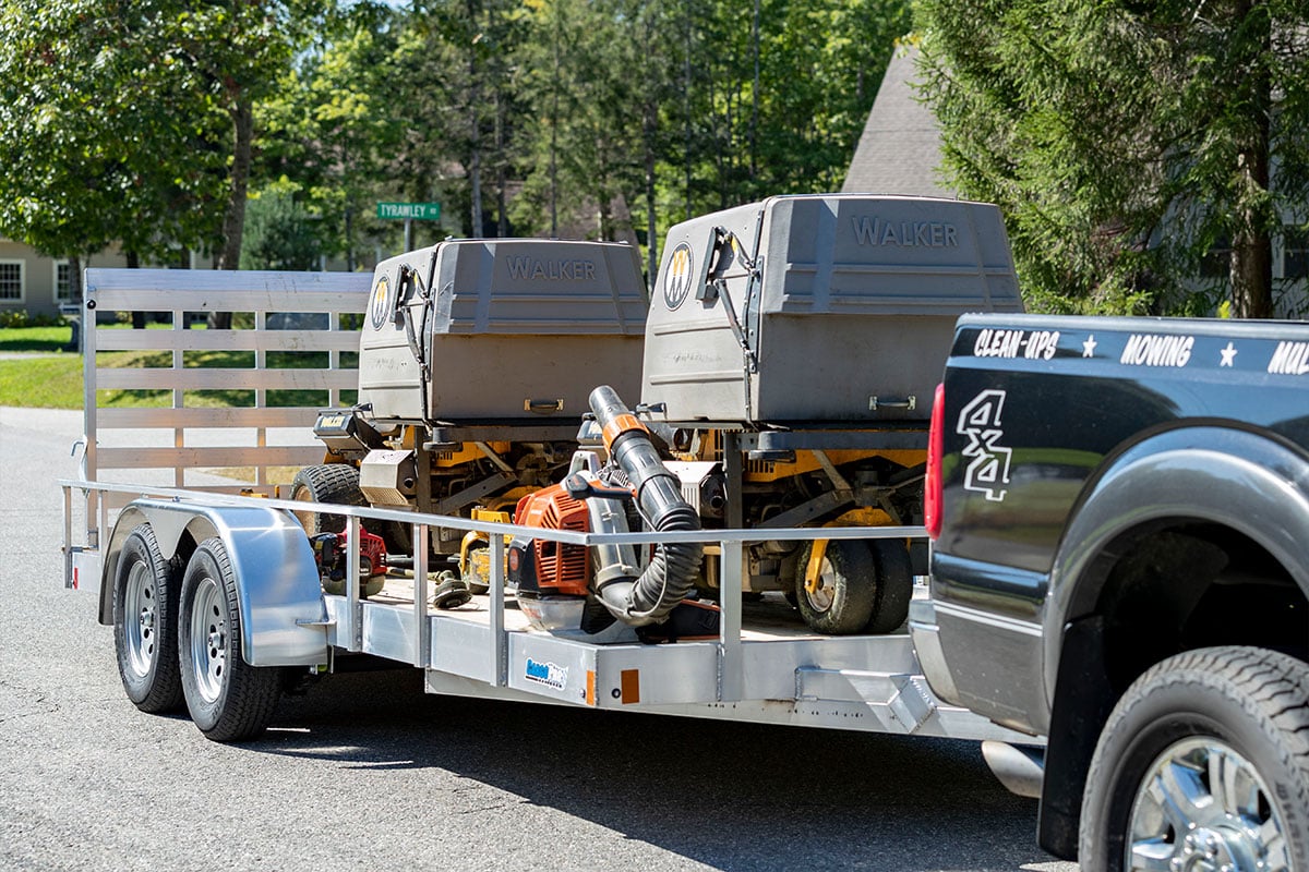 Open aluminum utility trailer by ALCOM loaded with lawn care equipment, parked at a jobsite. 