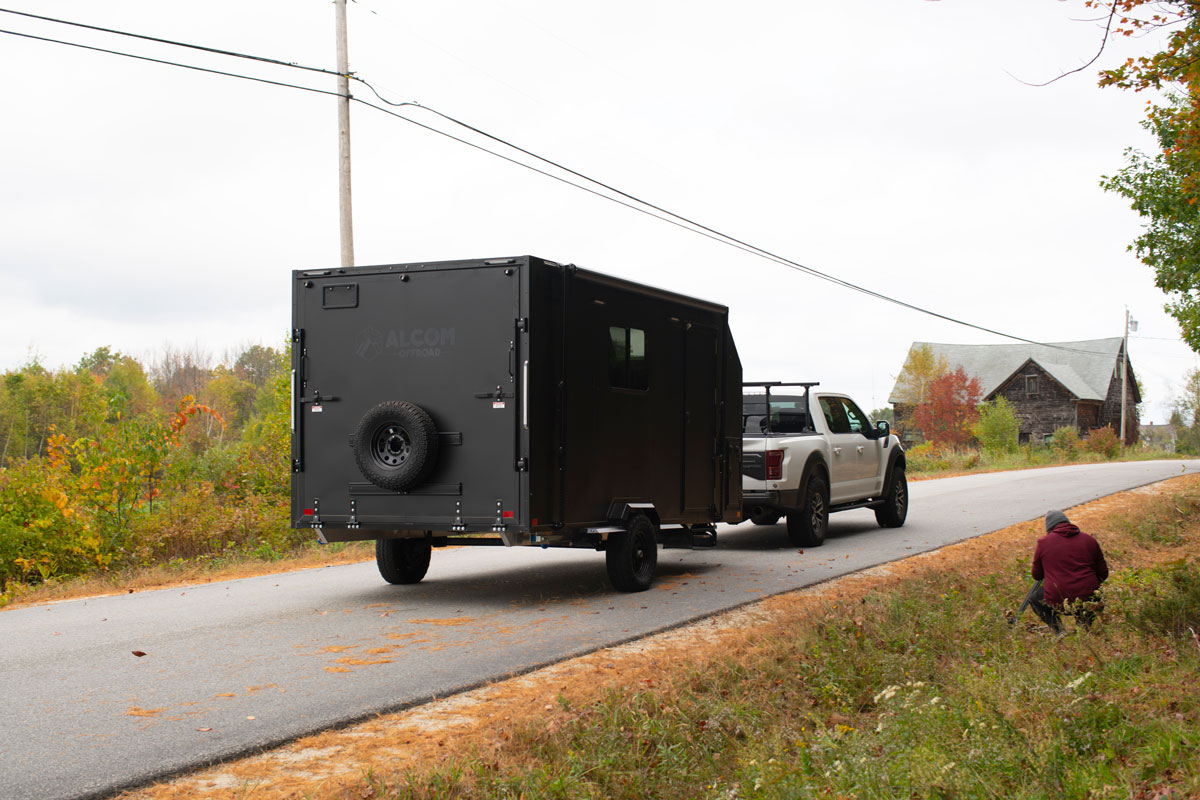 An AO Trailers black Caldera offroad trailer towed by a white pickup on a paved road