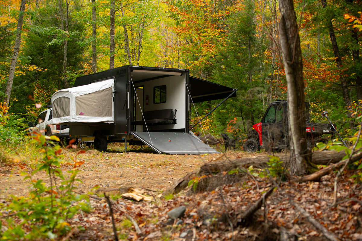 A black AO Trailers Caldera offroad camper and toy hauler set up at a wooded campsite with a dog playing nearby and UTV unloaded