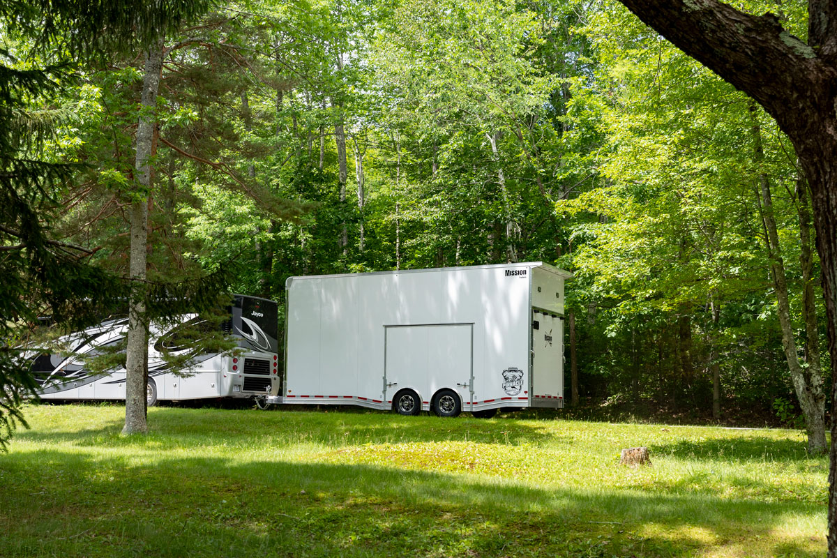 White enclosed Pinnacle Stacker car hauler trailer hitched to an RV driving through trees on a sunny day