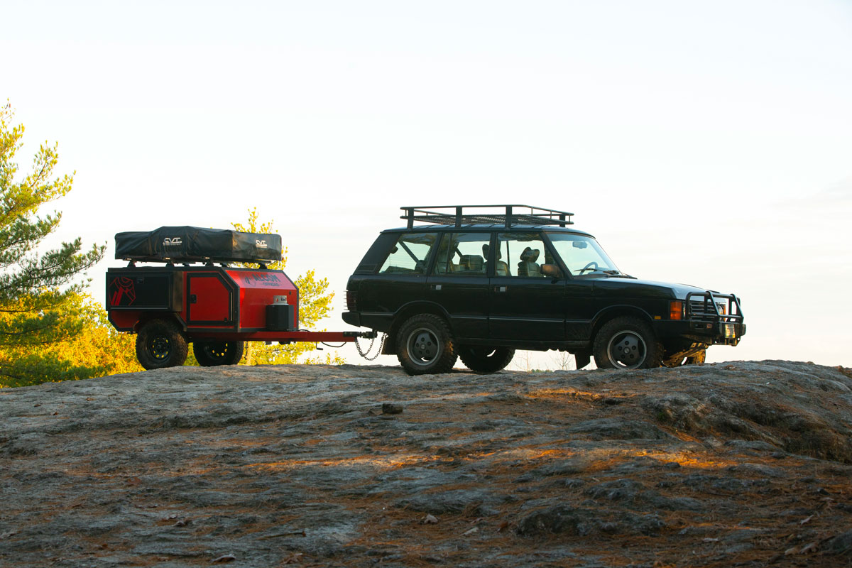 A lightweight red Spur offroad trailer by AO Trailers towed by an SUV to the top of a rocky hill overlooking the sunset
