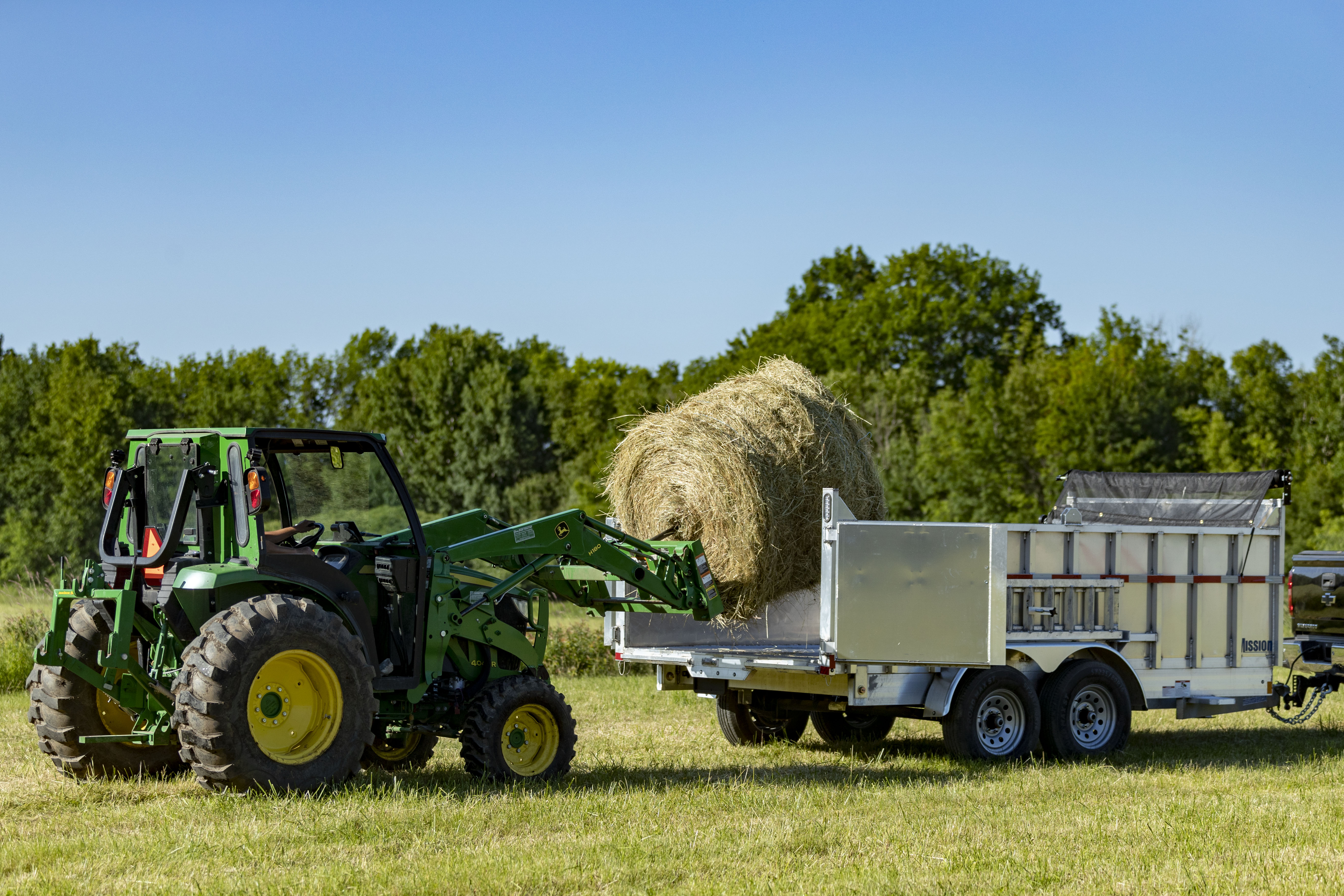 Loading a hay roll into an aluminum commercial dump trailer with a green farm tractor on a sunny day