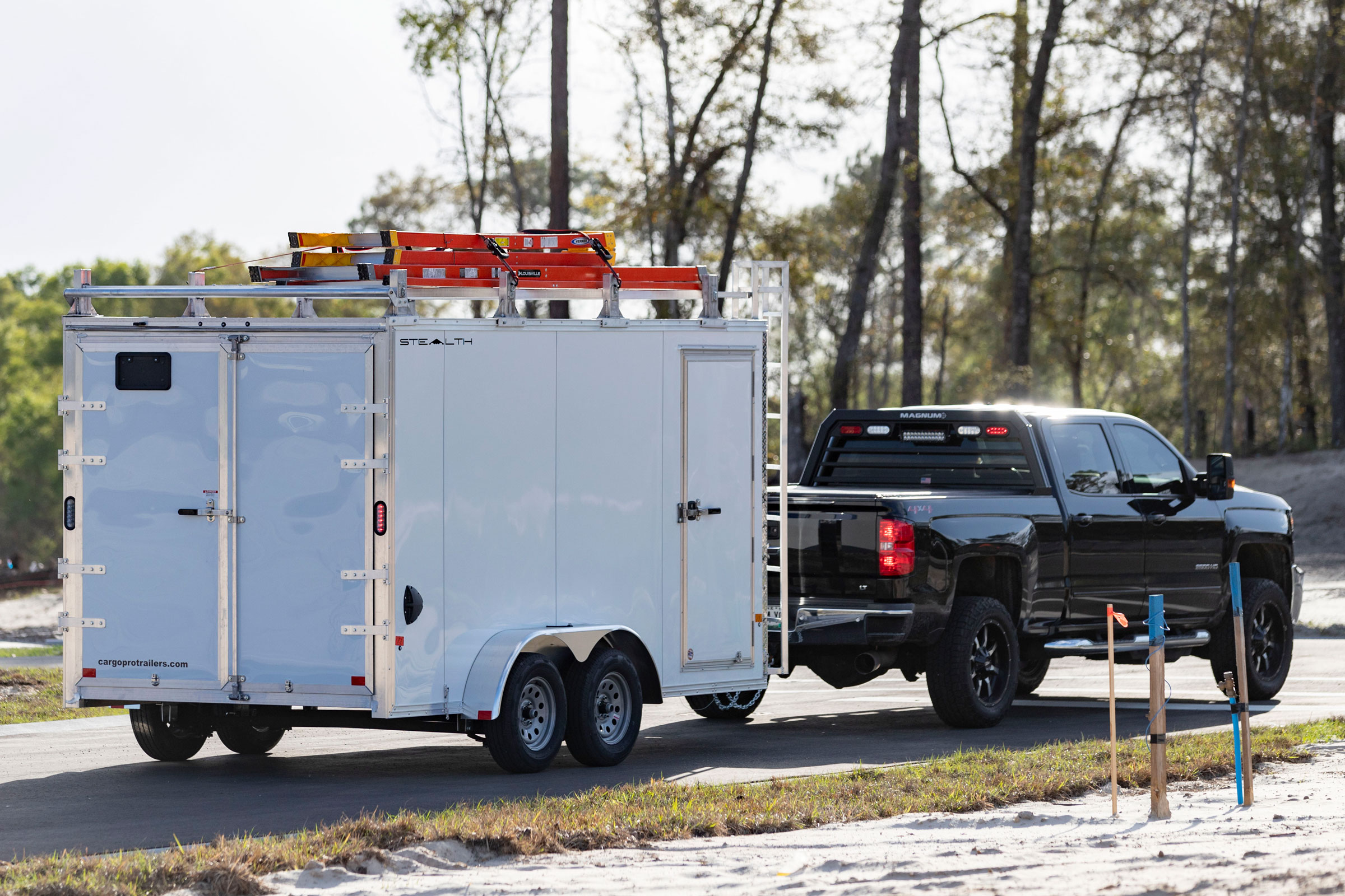 A white enclosed aluminum contractor trailer towed by a black pickup on a sunny road